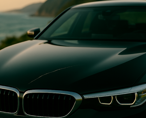 Black automotive paint on a sedan hood at dusk, with a noticeable scratch cutting across the glossy surface and reflecting ambient light.